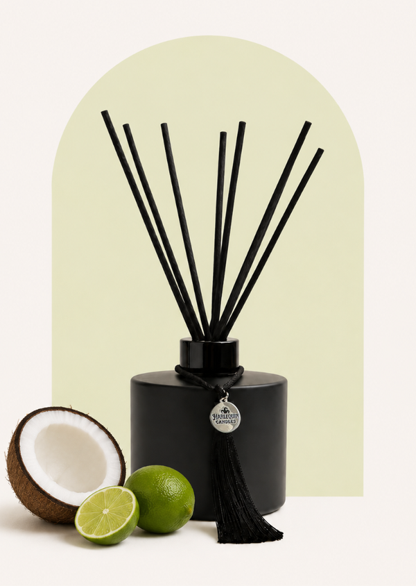 An Australian made Harlequin Candles black matte reed diffuser with a tropical coconut and lime scent called Coco Lime Sublime. The bottle is on a green background with sliced limes and cracked coconut.