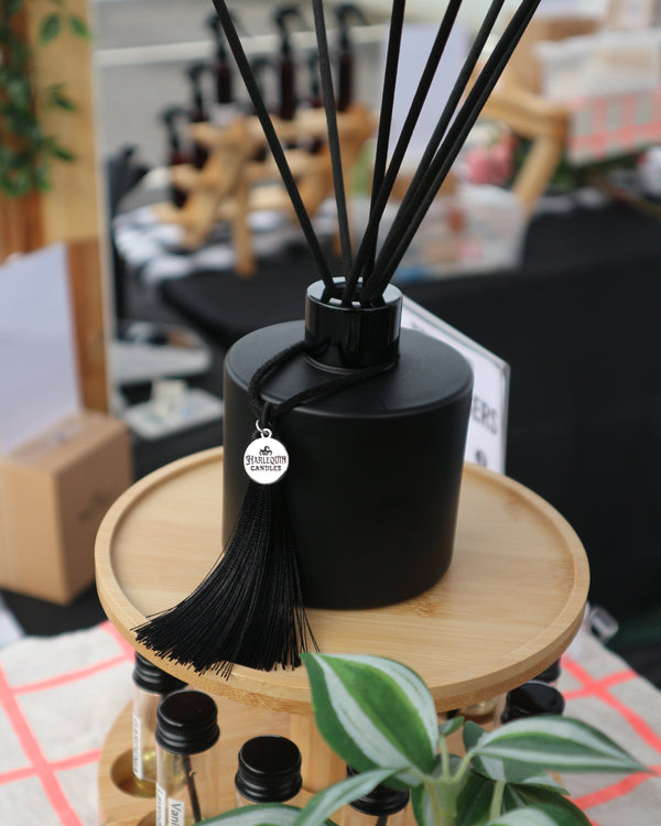 Black Harlequin Candles reed diffuser with fibre reeds and tassel displayed with fragrance testers at Bentleigh Sunday Market in Melbourne.