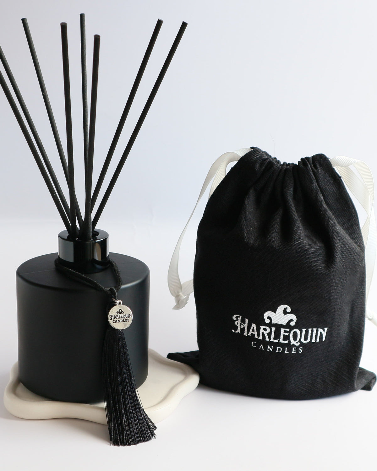 A sleek matte black Harlequin Candles reed diffuser bottle with black reeds, a black tassel charm, and a black drawstring gift bag featuring the white Harlequin Candles logo. The items are artfully arranged on a subtle white coaster against a clean white background, showcasing a luxury home fragrance product.
