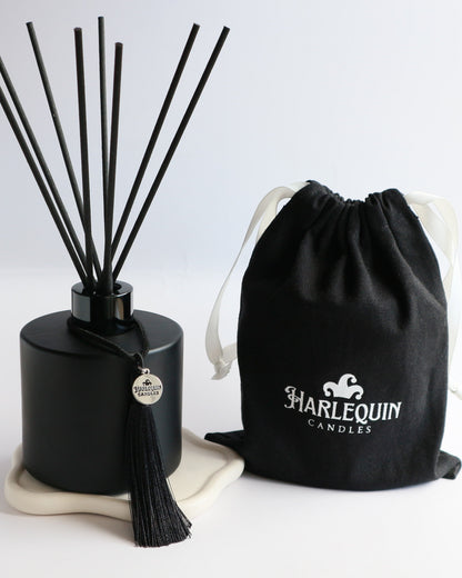 A sleek matte black Harlequin Candles reed diffuser bottle with black reeds, a black tassel charm, and a black drawstring gift bag featuring the white Harlequin Candles logo. The items are artfully arranged on a subtle white coaster against a clean white background, showcasing a luxury home fragrance product.