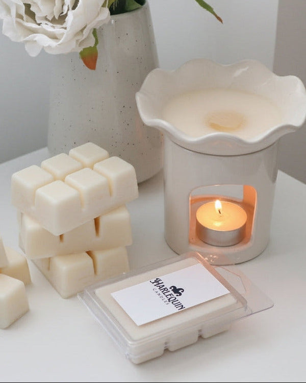 A stack of Harlequin Candles premium soy wax melts next to a white ceramic wax warmer with a lit tealight candle and melted wax. A clear plastic clamshell of wax melts with the Harlequin Candles label is in the foreground, showcasing eco-friendly home fragrance.