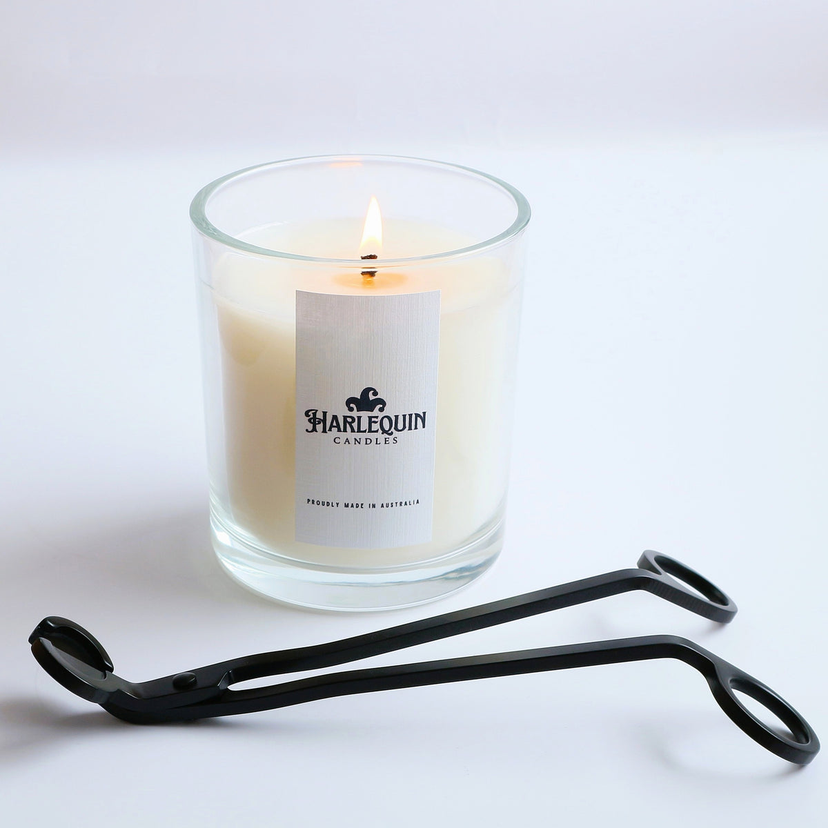 A lit Harlequin Candles soy candle in a clear glass jar with a white label reading 'PROUDLY MADE IN AUSTRALIA', accompanied by a black candle wick trimmer, all on a clean white background. Showcases a premium Australian-made candle and essential wick trimmer tool.