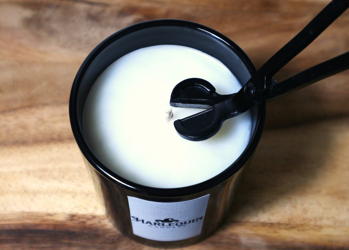 Close-up of a black candle wick trimmer actively trimming the wick of a Harlequin Candles soy candle in a black jar. Illustrates essential candle care for a clean and efficient burn, promoting longer candle life.