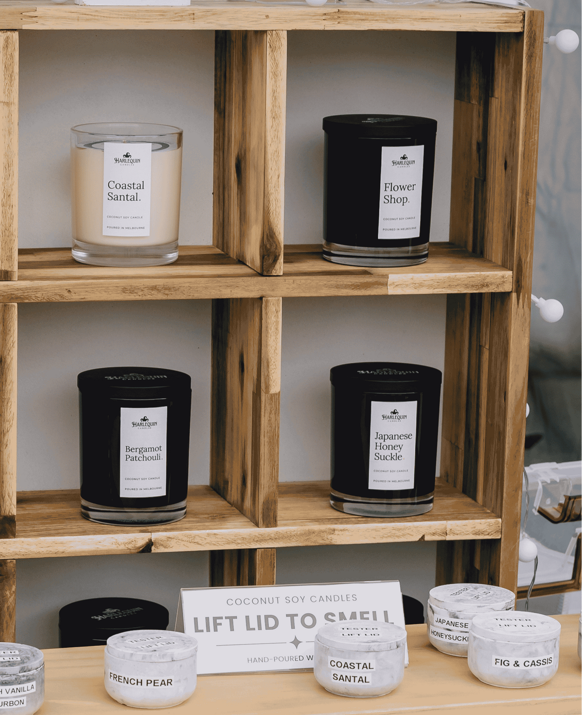 A wooden display shelf showcasing a collection of scented soy candles from Coconut Soy Candles, featuring various sizes and scents like Fig & Cassis, Coastal Santal, and Flower Shop in glass and metal jars.