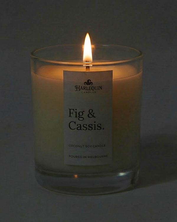 A lit Fig & Cassis coconut soy candle by Harlequin Candles in a clear glass jar. The warm flame illuminates the minimalist white label, which features the brand's logo and the text "Poured in Melbourne" against a soft, dark background.