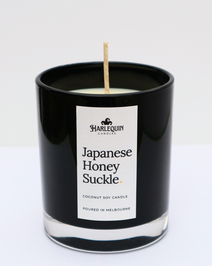 A sleek black glass jar containing a Japanese Honeysuckle coconut soy candle by Harlequin Candles. The minimalist white label features bold black text and the brand's logo, with the text "Poured in Melbourne" at the bottom against a clean, bright background.