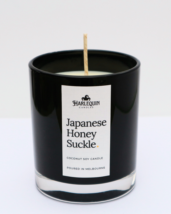 A sleek black glass jar containing a Japanese Honeysuckle coconut soy candle by Harlequin Candles. The minimalist white label features bold black text and the brand's logo, with the text "Poured in Melbourne" at the bottom against a clean, bright background.