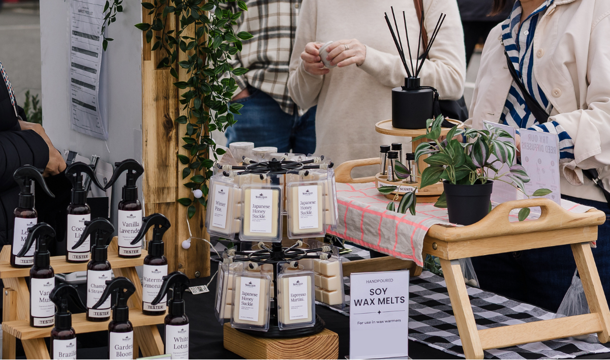 A market stall displaying room mist products, including spray bottles and wax melts, with shoppers browsing nearby.